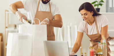 une personne emballe des boîtes à lunch dans un sac blanc, et une autre personne travaille sur un ordinateur portable.